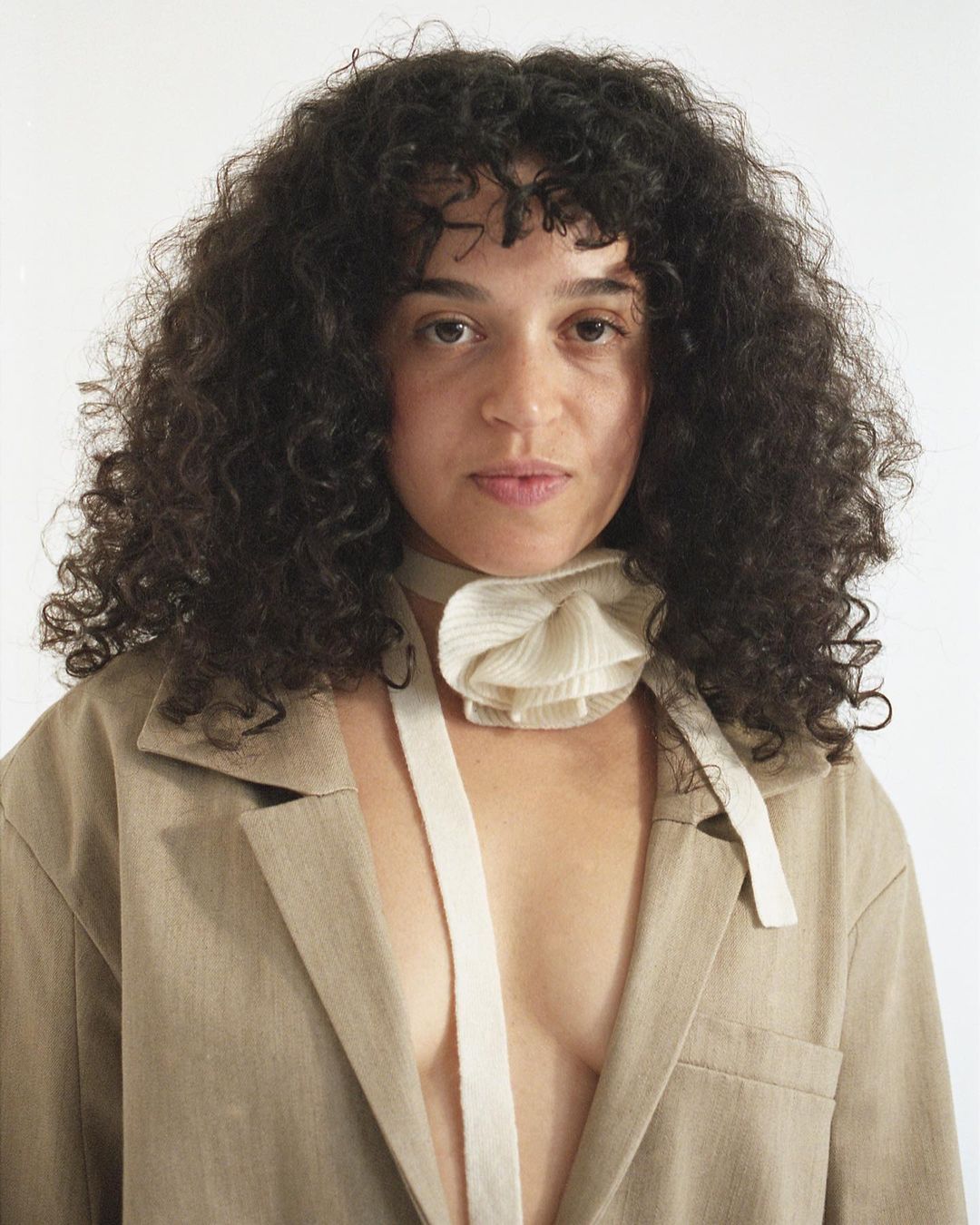 Portrait of a woman with dark curly hair wearing a beige blazer and a decorative fabric scarf in a studio setting against a white backdrop.