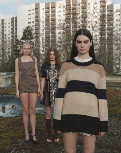 Three female models in a minimalistic outdoor fashion shoot, featuring a high-rise apartment building and overcast sky.