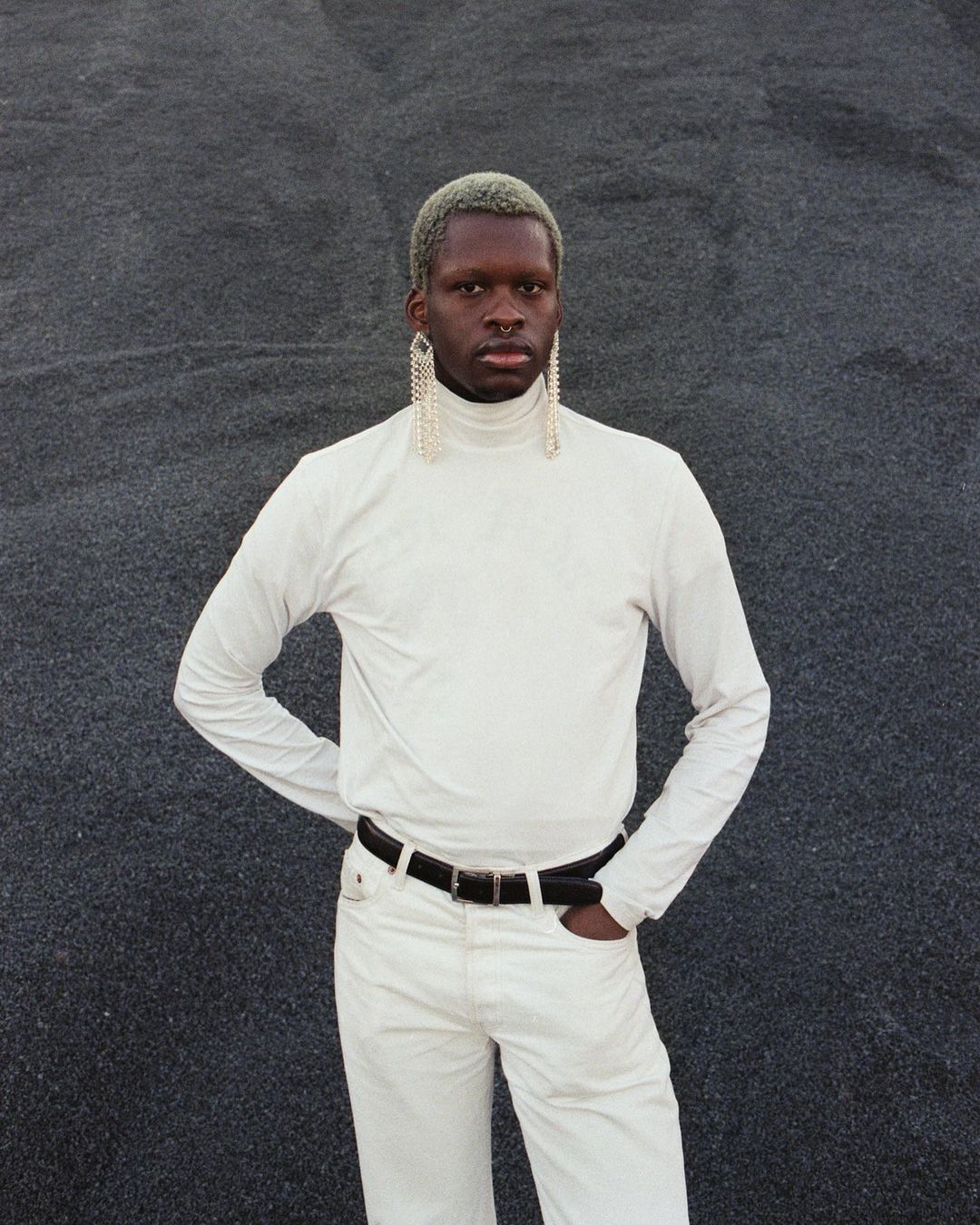 A person with platinum hair and dramatic earrings poses against a grey backdrop, wearing a white turtleneck and matching pants.