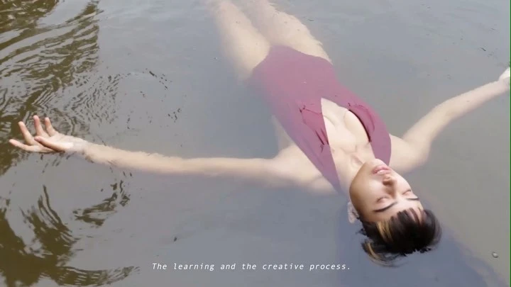 Woman in a plum-colored one-piece swimsuit floats in a body of murky water with her arms outstretched in an outdoor location.