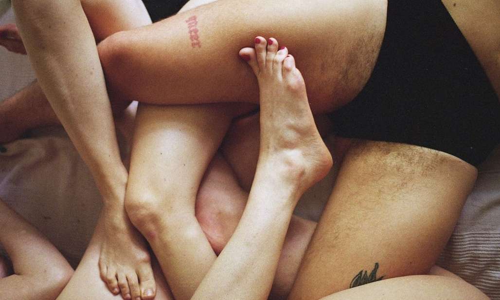 Close-up of intertwined legs and feet of three people lying on striped bedding, with visible tattoos on skin.