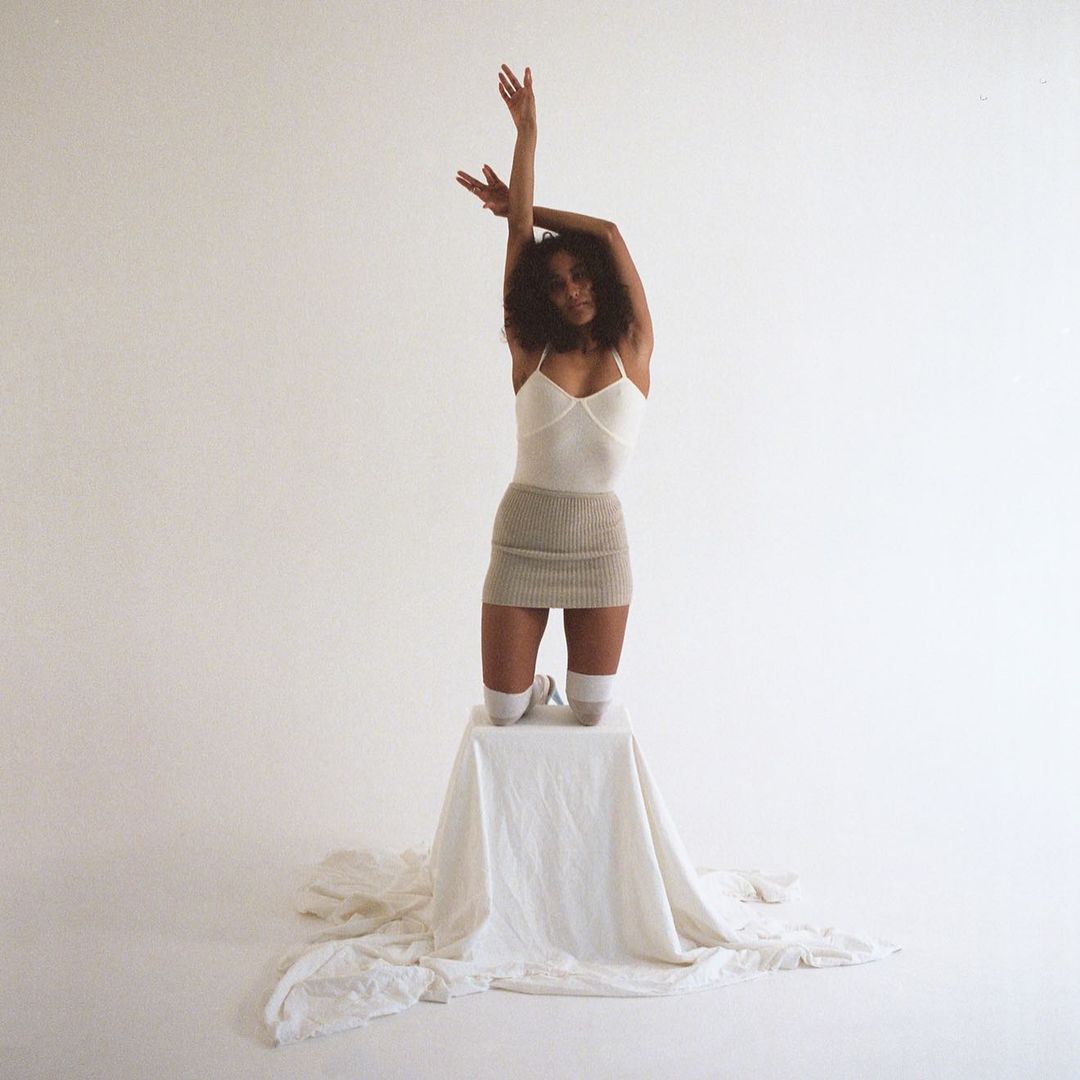 Woman in a tan knit skirt and white top poses on a white cube in a bright studio, arms raised, with a minimalist fashion aesthetic.