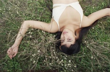 Woman lying flat on her back amidst green grass and small white wildflowers, wearing a white wrap-style bikini top.