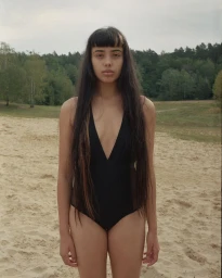 Portrait of a woman with long dark hair and bangs wearing a black swimsuit standing on a sandy beach.