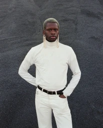 Black man wearing a white turtleneck and white pants with long crystal earrings against dark gravel by Celine Yasemin