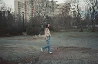 Young woman walking across a paved open area wearing flared blue jeans and a light top, with apartment buildings in the background.