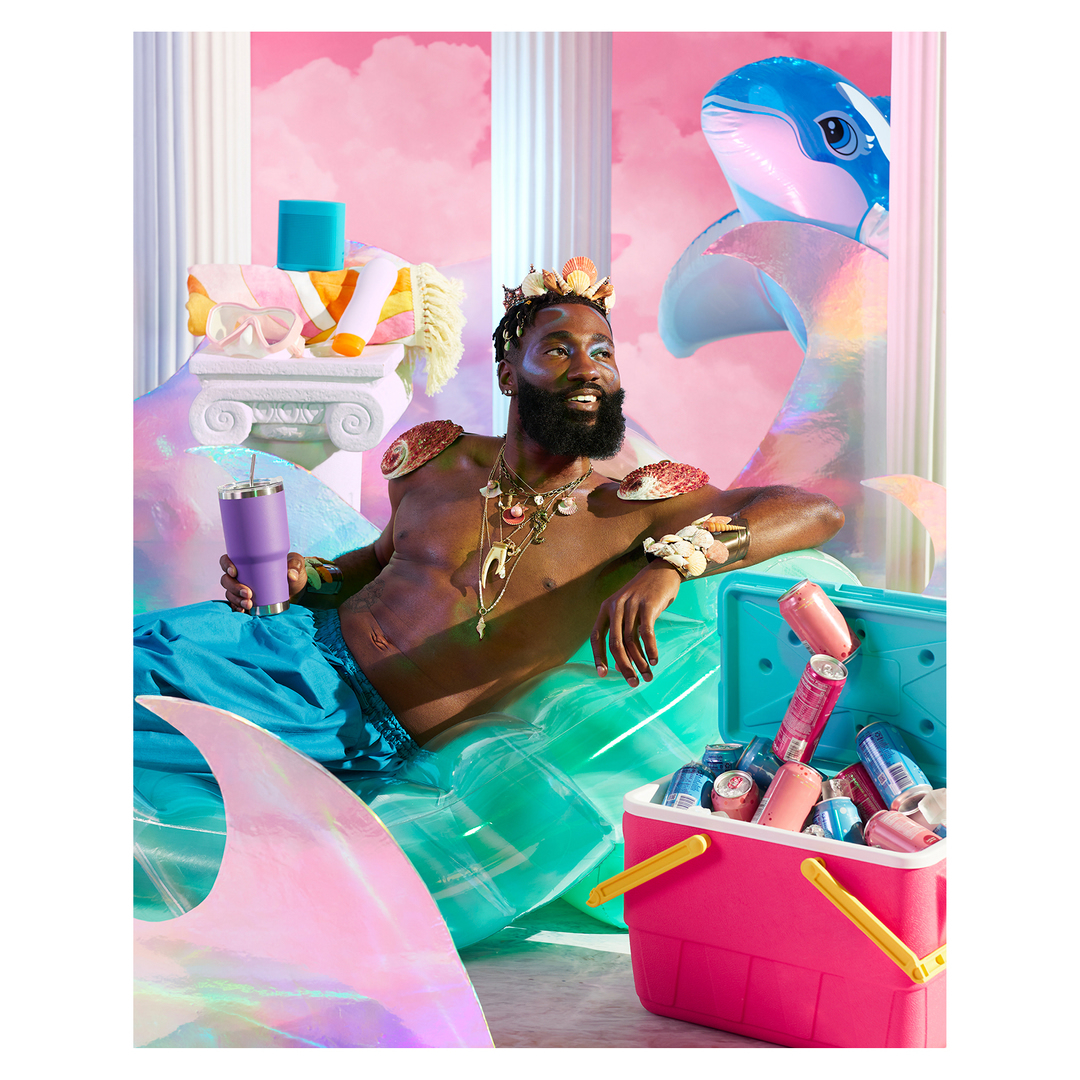 Man with a beard wearing seashells and turquoise shorts, holding a purple cup, reclining in a surreal scene with inflatable whale.