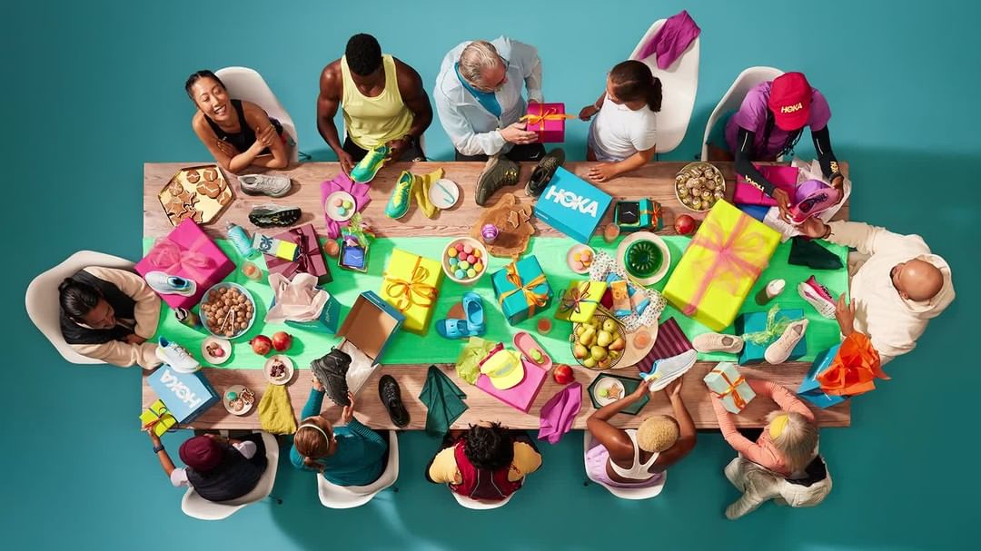 Overhead view of diverse group seated at table covered with running shoes, wrapped gifts, and snacks against a turquoise background.