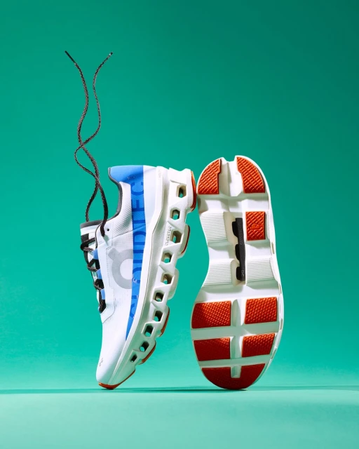 Studio shot of white and blue athletic shoes, one on its side showing the sole with orange and black details, on a teal background.