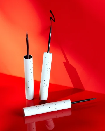 Three white speckled eyeliner pens with black applicator brushes positioned against a vivid red studio background