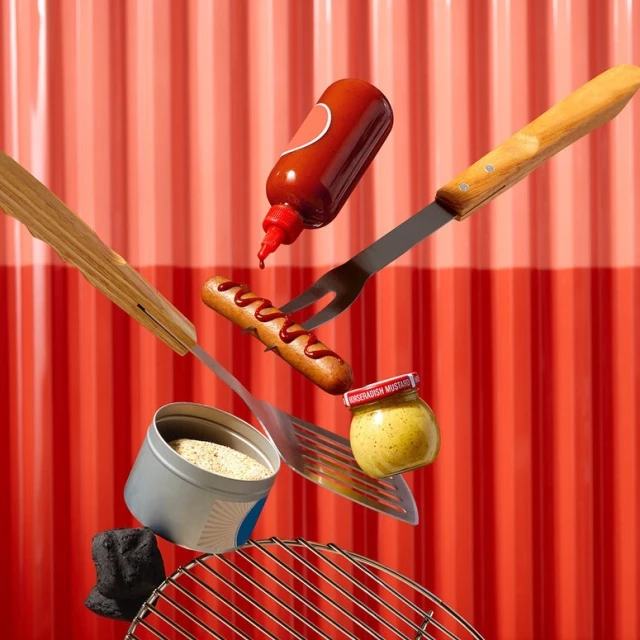 Still life of sausage on a fork, ketchup, mustard, spatula, grill, and charcoal, arranged against a red corrugated metal background.