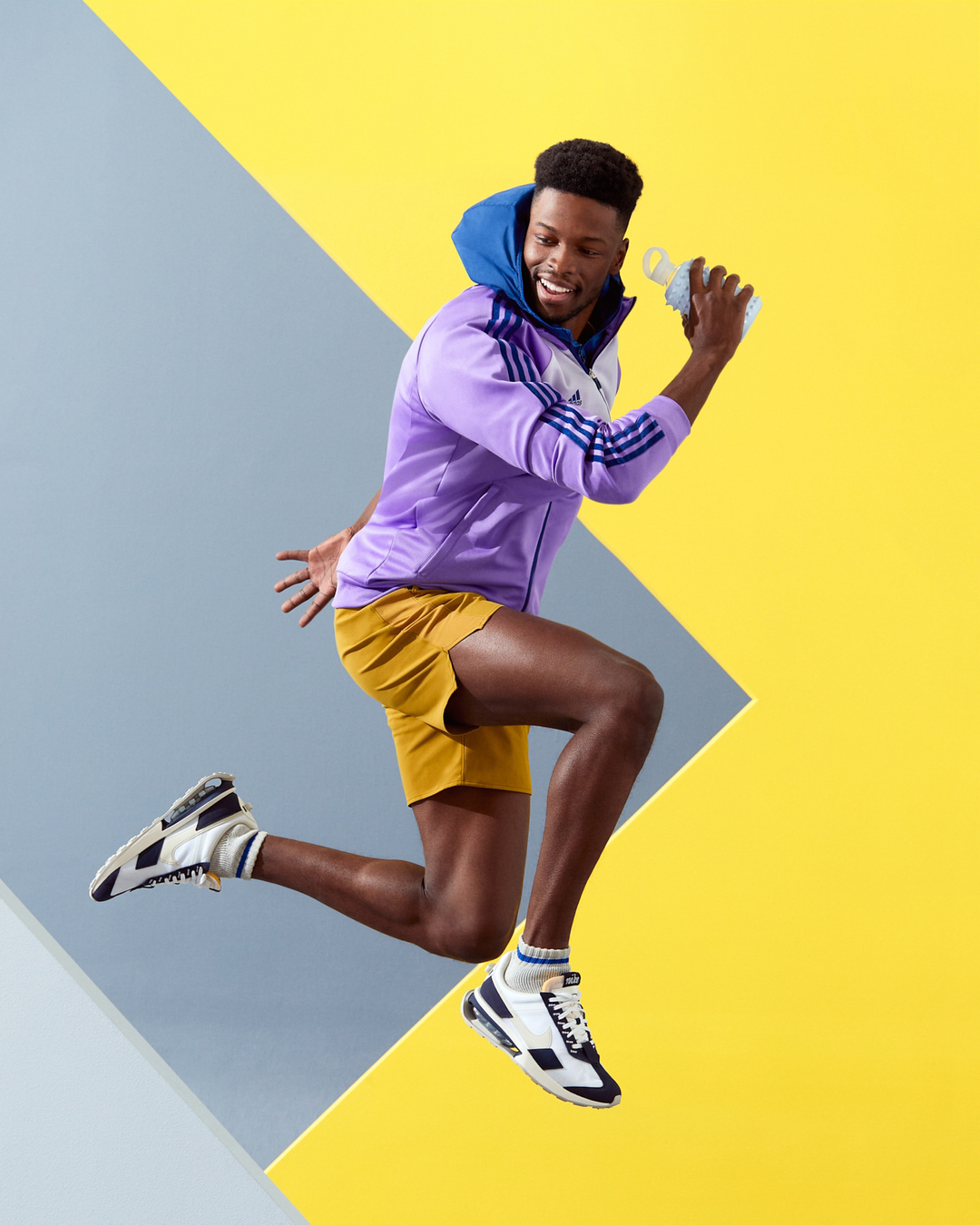 Athletic African-American man in sportswear jumping in front of a two-tone yellow and gray background while holding a water bottle.