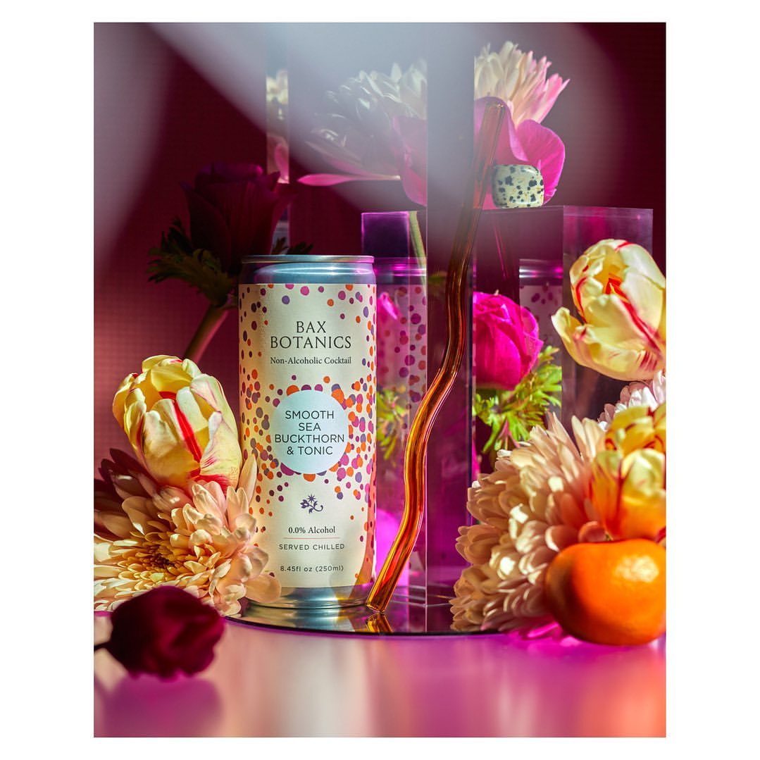 An aluminum can of Bax Botanics cocktail surrounded by colorful flowers and clear geometric shapes against a dark pink background.