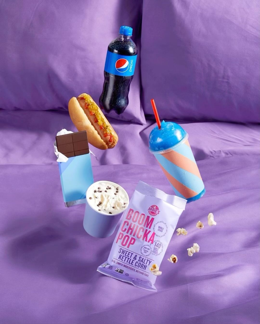 A tempting still life with a hot dog, Pepsi, chocolate, popcorn, and a slushy, all arranged on a vibrant purple sheet.