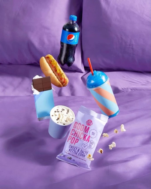 A tempting still life with a hot dog, Pepsi, chocolate, popcorn, and a slushy, all arranged on a vibrant purple sheet.