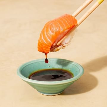 Salmon sushi nigiri held with chopsticks dripping sauce into a green bowl against a light peach backdrop in a food and beverage setting.