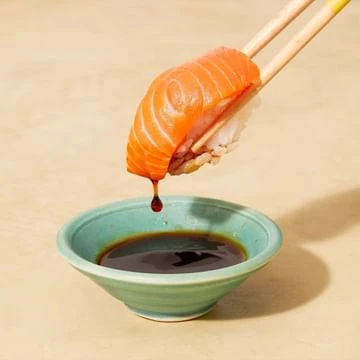 Salmon sushi nigiri held with chopsticks dripping sauce into a green bowl against a light peach backdrop in a food and beverage setting.