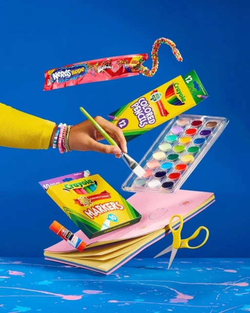 A hand holds a paintbrush among floating school and art supplies, including candy, colored pencils, markers, a watercolor set, and scissors.