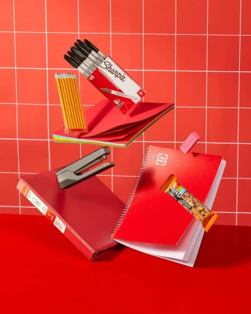Stationery and school supplies including notebooks, pens, pencils, stapler, and a granola bar float against a red grid background.