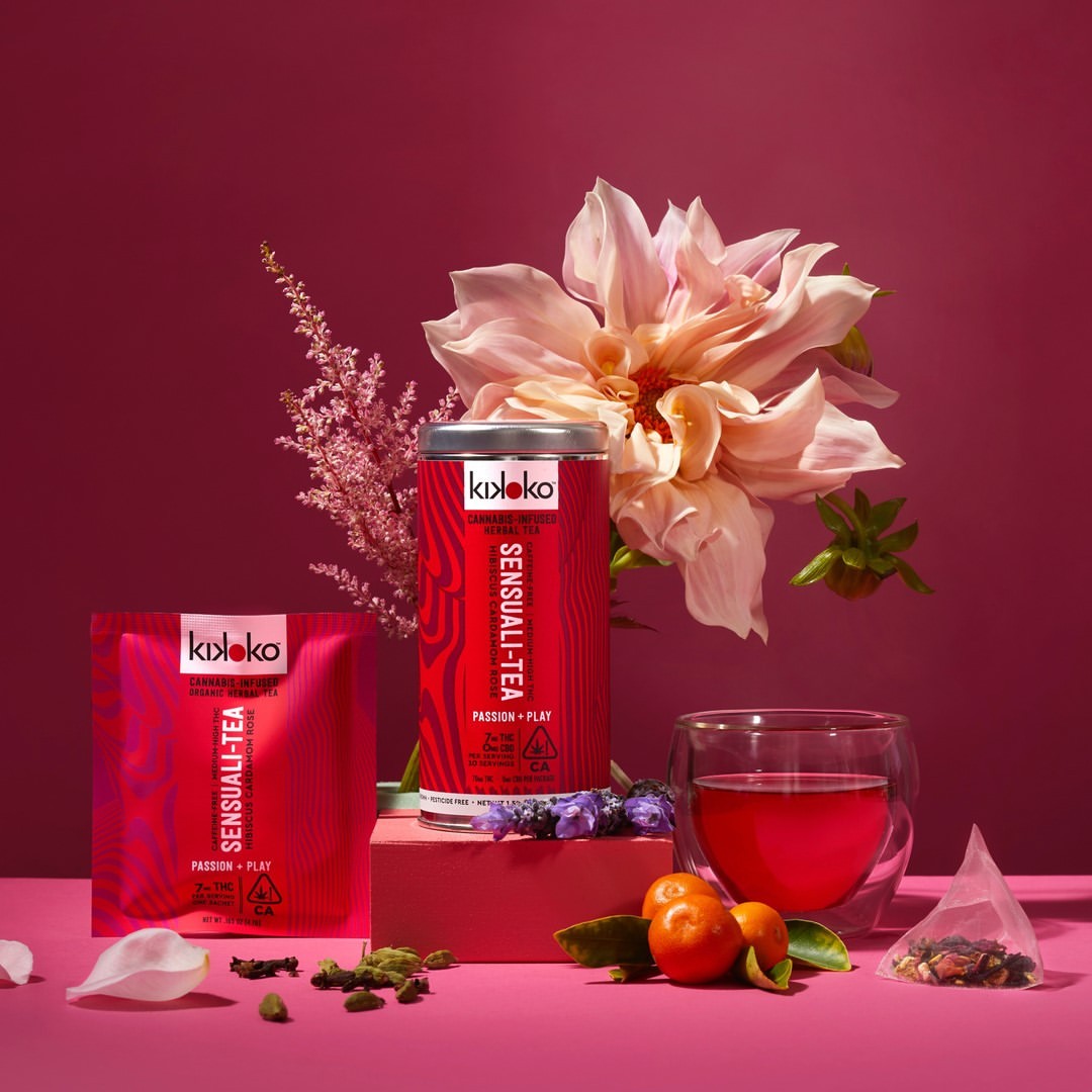 Kikoko Sensuali-Tea products on pink background with pink flowers, a clear tea cup, and loose tea; eye-level, medium shot.