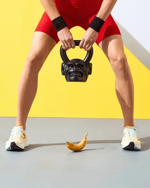 Fitness-focused shot of an athlete holding a kettlebell with a gorilla face, positioned above a ripe banana on a gray and yellow backdrop.