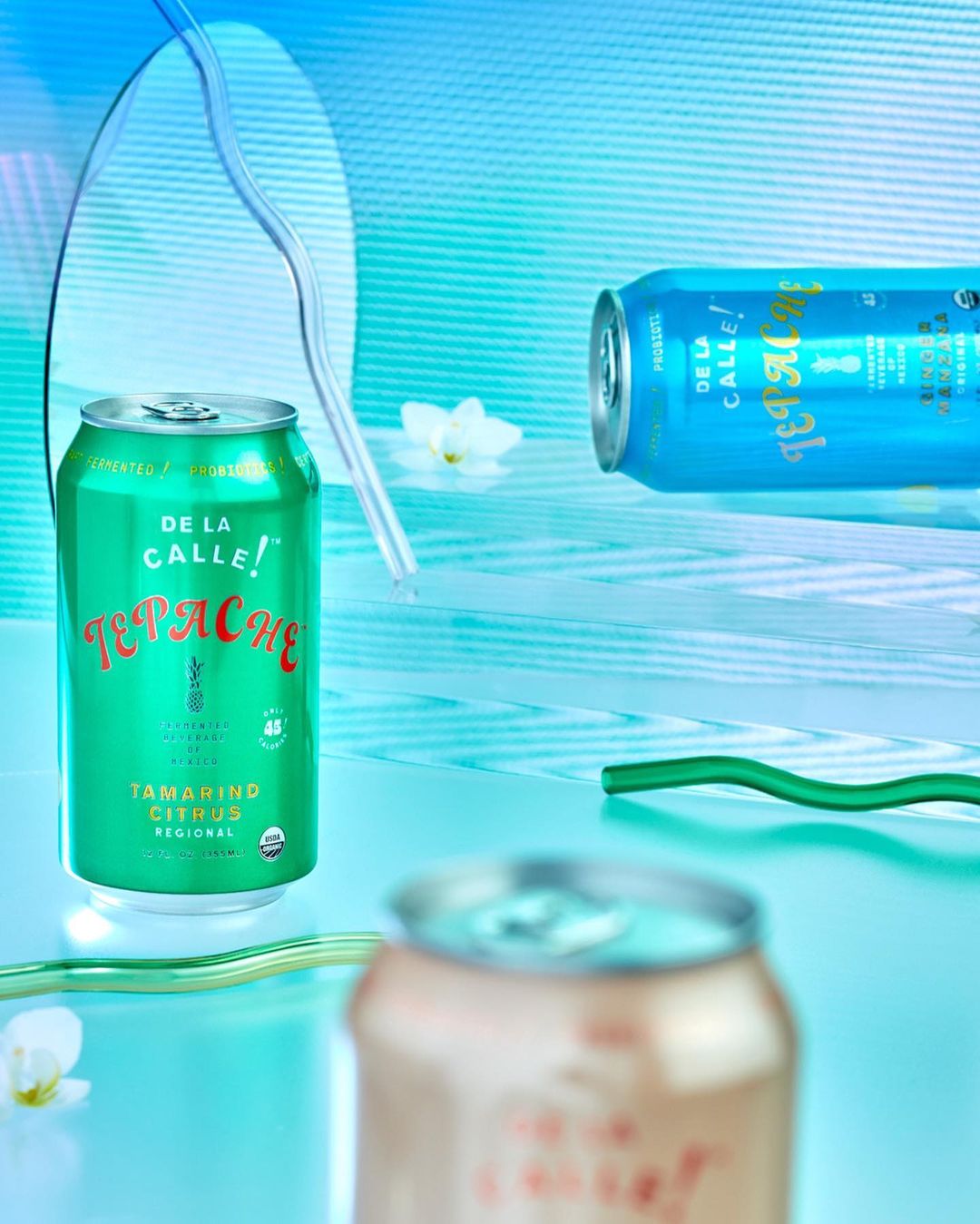 Three cans of De La Calle Tepache flavored drinks are displayed with bent straws and white flower blossoms against a light teal backdrop.