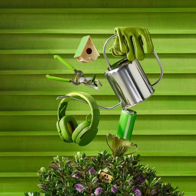 Still life of a watering can, headphones, trophy, gardening tools, birdhouse, and a battery on top of a plant with green background.