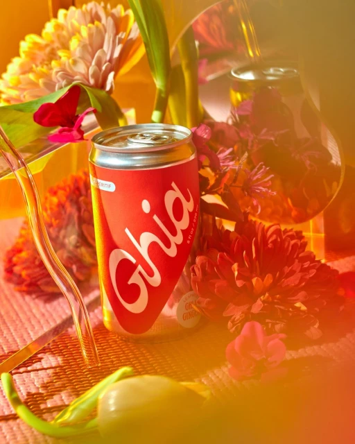 A single can of Ghia stands beside colorful flowers, set against a softly textured background, creating a vibrant, eye-catching beverage still life.