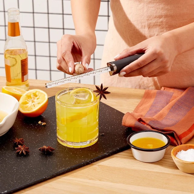 Cocktail preparation featuring a woman grating nutmeg over a yellow drink garnished with star anise and lemon on a black serving board.