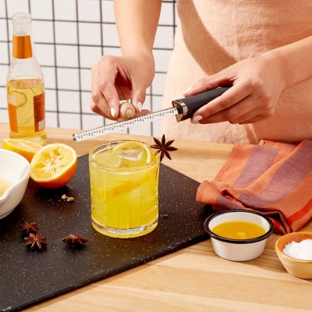 Cocktail preparation featuring a woman grating nutmeg over a yellow drink garnished with star anise and lemon on a black serving board.