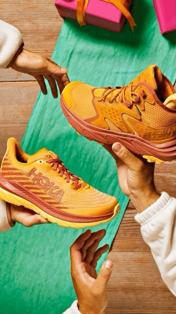 Two Hoka running shoes are presented by human hands against a green cloth and wood background with pink wrapped gifts above.