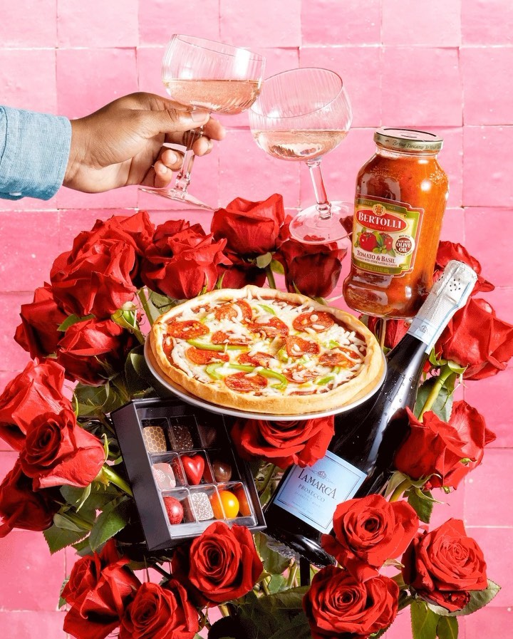 Celebration still life with pizza, wine, roses, and chocolates for a romantic food and beverage scene on pink backdrop.