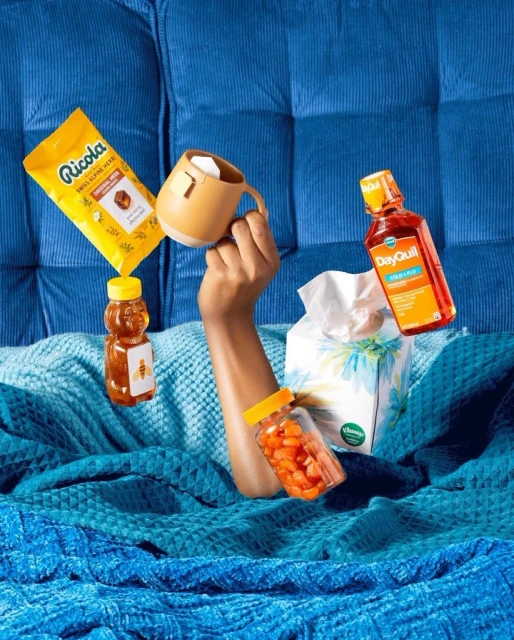 Hand raises mug with cold remedy products: cough drops, honey, medicine, and tissues on a blue corduroy couch and blanket.