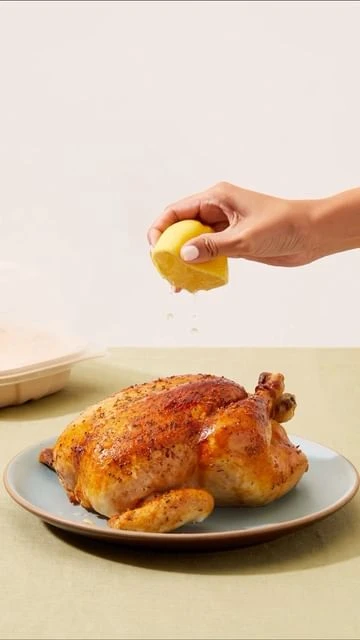 Overhead shot of a roasted chicken on a blue plate being squeezed with a lemon by a female hand against a neutral background.
