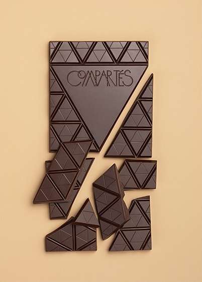 Dark chocolate bar broken into triangular pieces on a tan background with triangular pattern and the brand name Compartes. 