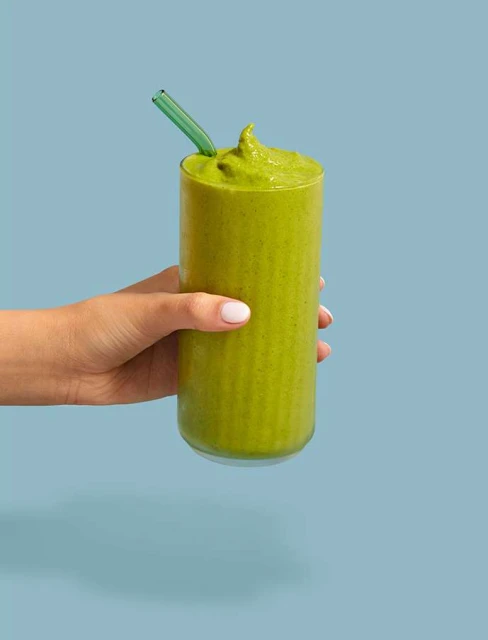 A hand holds a tall glass filled with a green smoothie and a green straw against a light blue background in a studio shot.