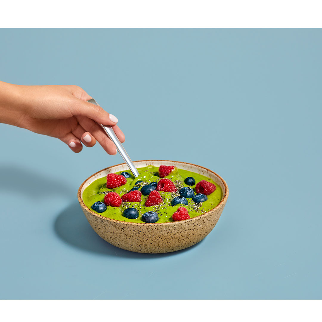 Close-up of a hand using a spoon to eat a green smoothie bowl topped with raspberries and blueberries on a blue background.