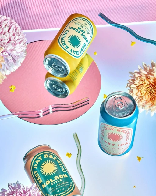 Three cans of Best Day Brewing beer in different colors, with colorful straws and floral elements, against a gradient background.