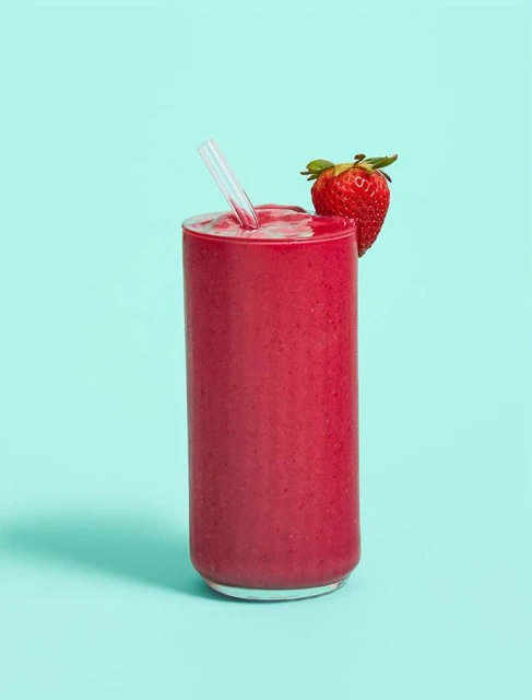 Eye-level shot of a tall glass filled with a vibrant red smoothie, garnished with a fresh strawberry and a clear straw against teal background.