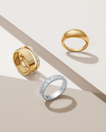 Three rings including two gold bands and one silver textured band on geometric beige surfaces with soft shadows.