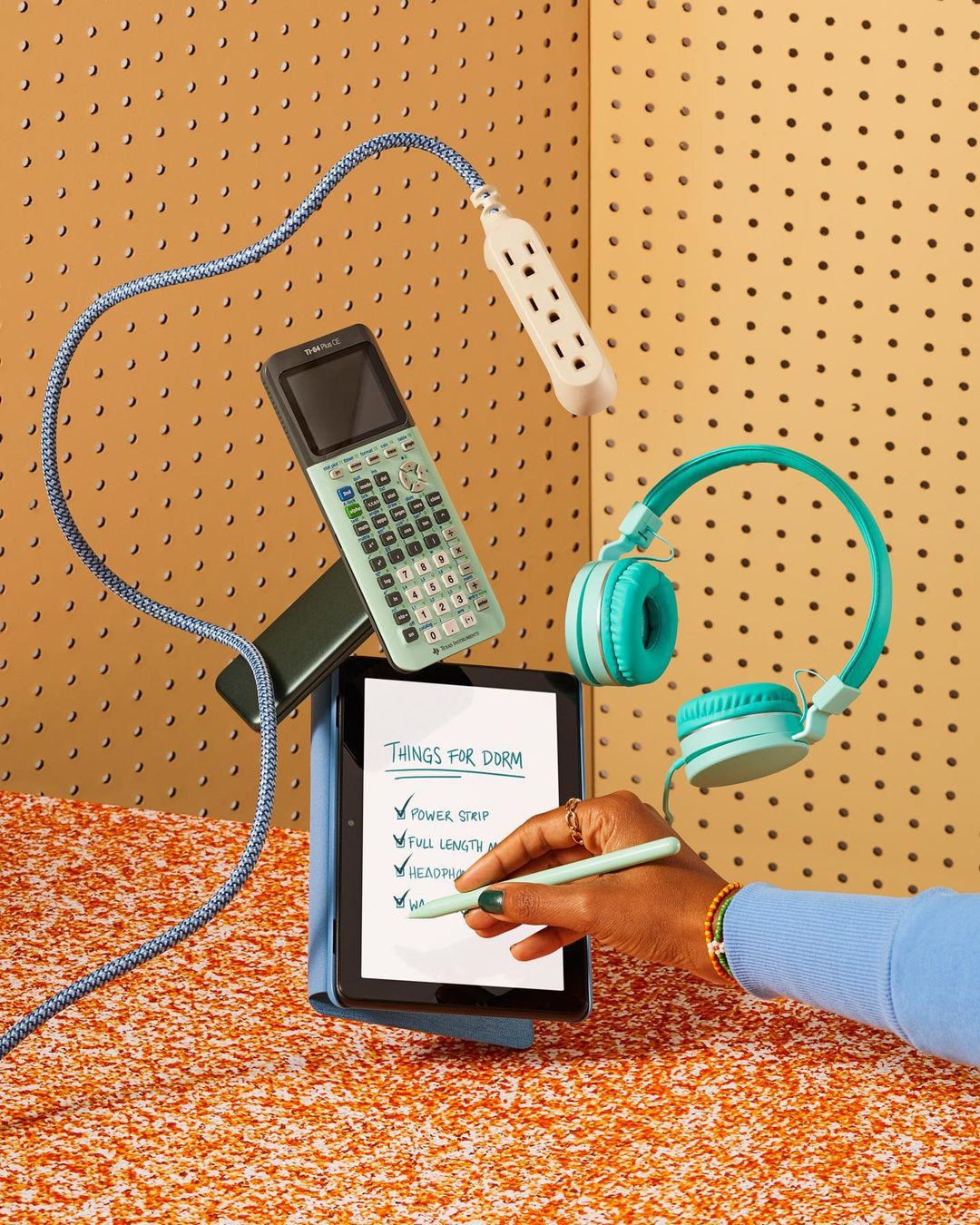 Flat lay photo of electronic devices and school supplies: tablet, calculator, headphones, power strip, pen, on an orange surface.