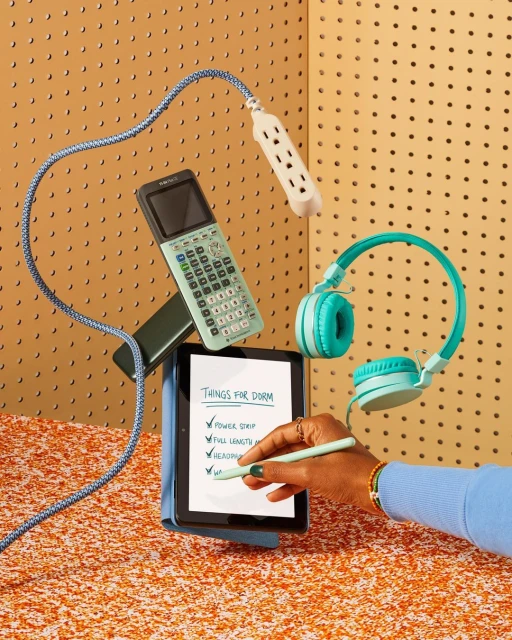 Flat lay photo of electronic devices and school supplies: tablet, calculator, headphones, power strip, pen, on an orange surface.