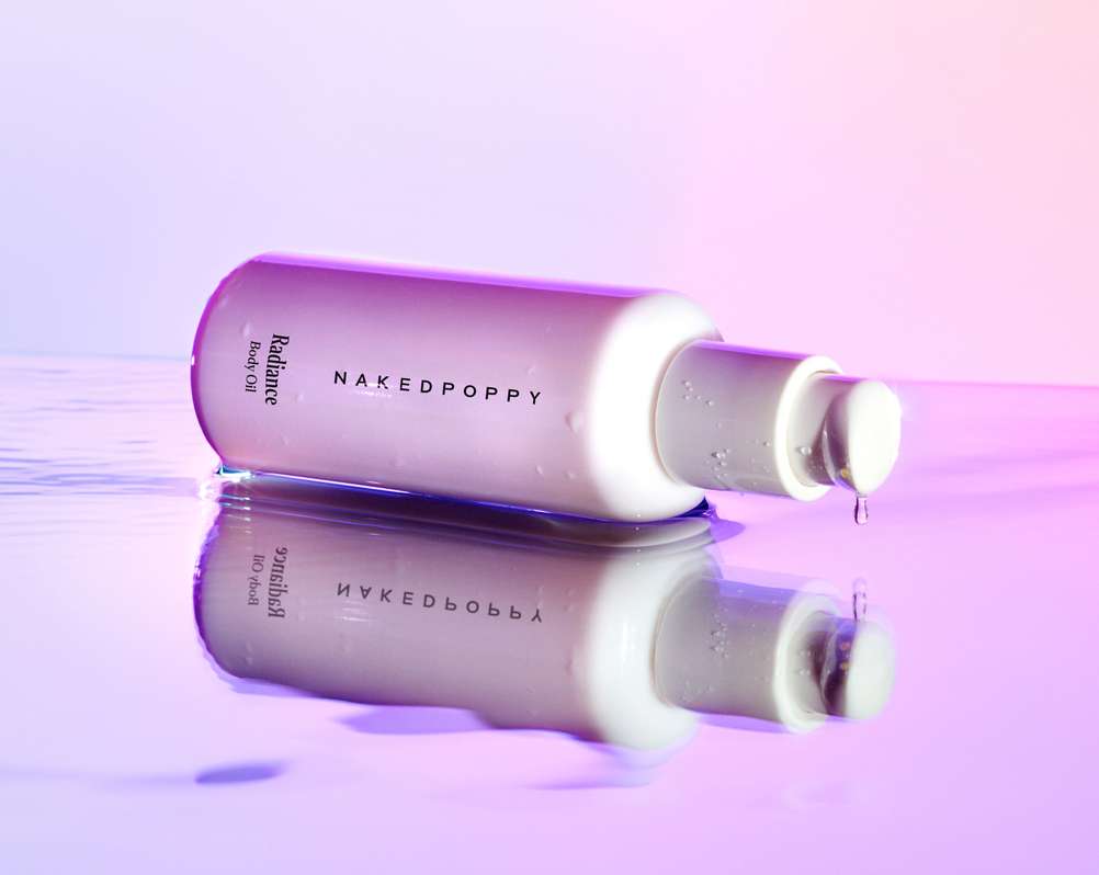 Naked Poppy radiance body oil bottle with pump, reflected in water with a pink and purple gradient background and water droplet falling.