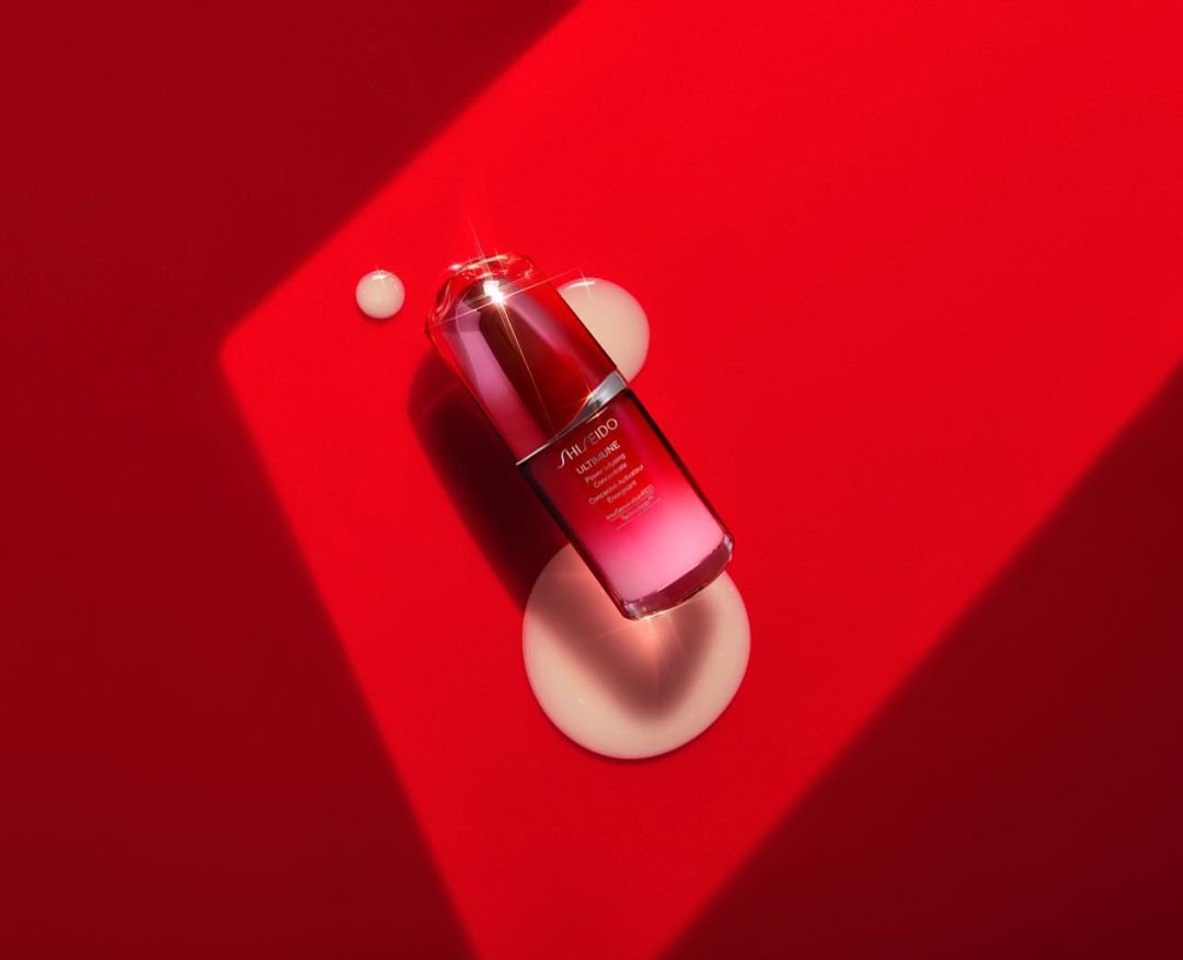Product shot of a red and clear Shiseido Ultimune serum bottle on a vibrant red surface with cream-colored cosmetic drops nearby.