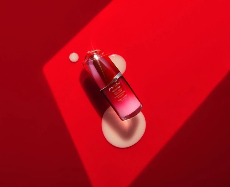 Product shot of a red and clear Shiseido Ultimune serum bottle on a vibrant red surface with cream-colored cosmetic drops nearby.