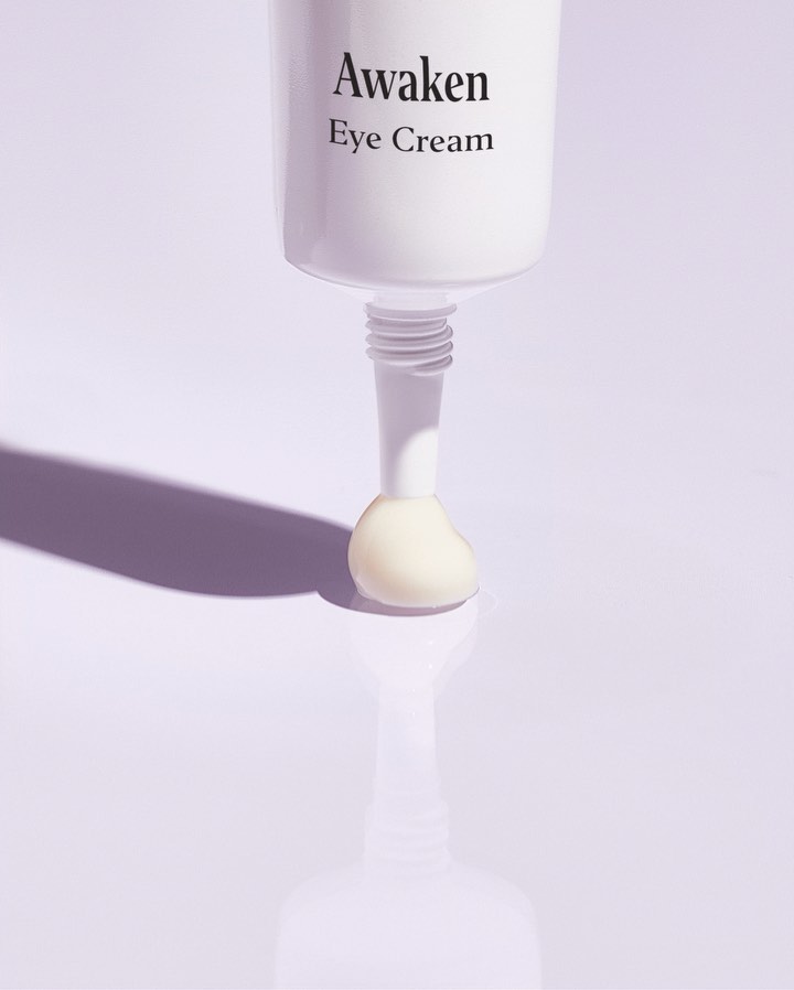 Close-up of white tube of eye cream, displaying squeeze-out of white cream on a light purple background with shadow and reflection.