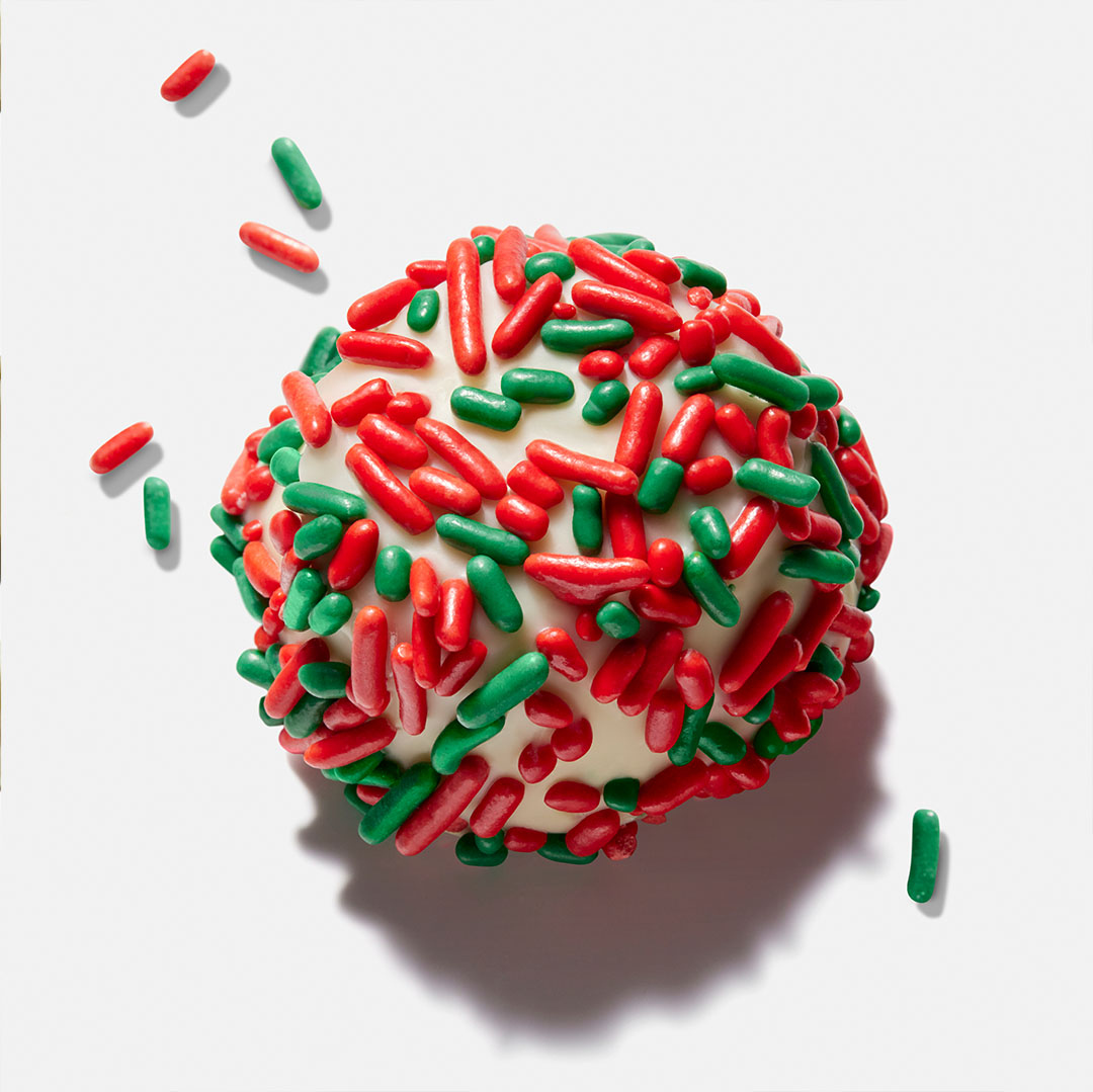 Close-up of a frosted cake ball covered in red and green sprinkles, set on a white surface and casting a soft shadow.