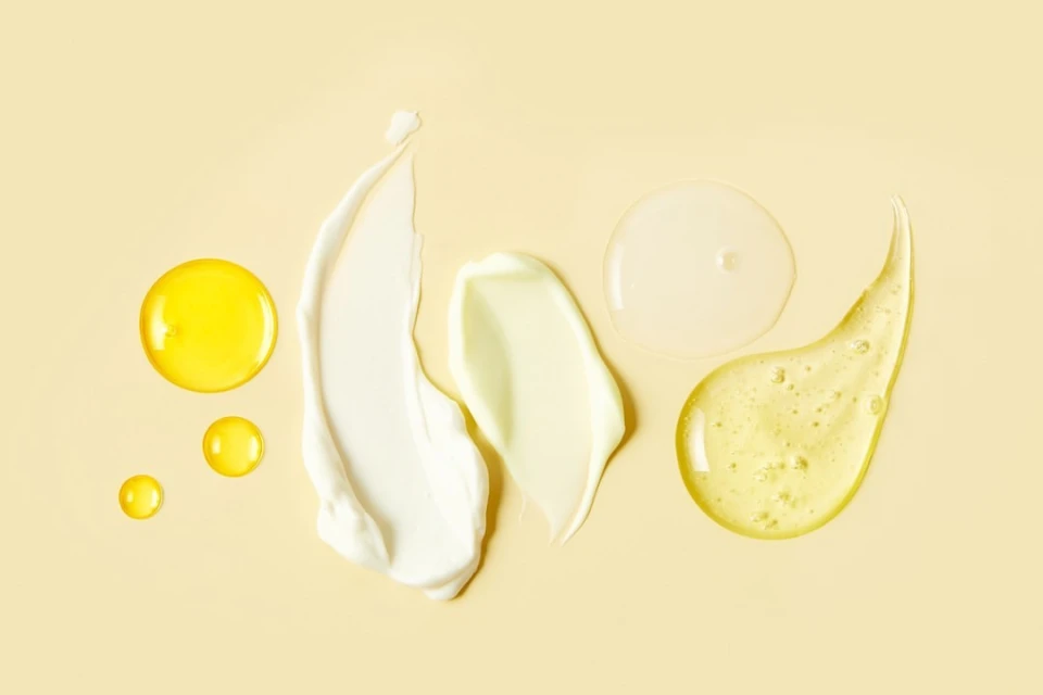 Cosmetic products including oil and creams displayed with various textures against a light yellow background in a flat lay composition.