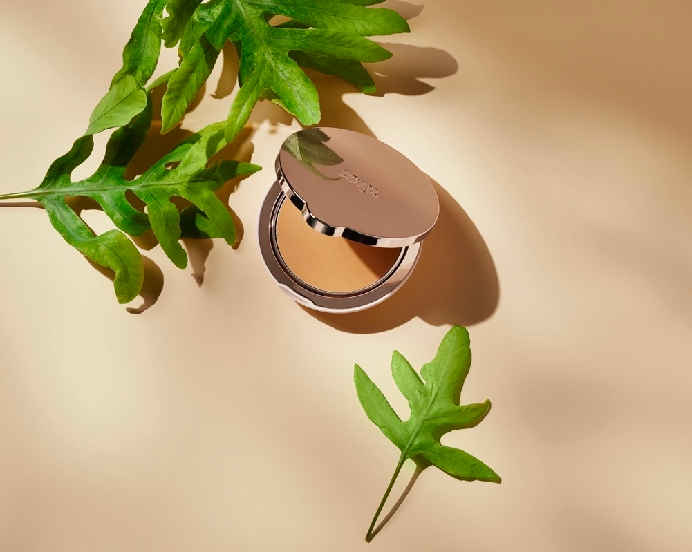 Powder foundation compact case with green leaves, on a beige background. Beauty product, natural light, product photography.