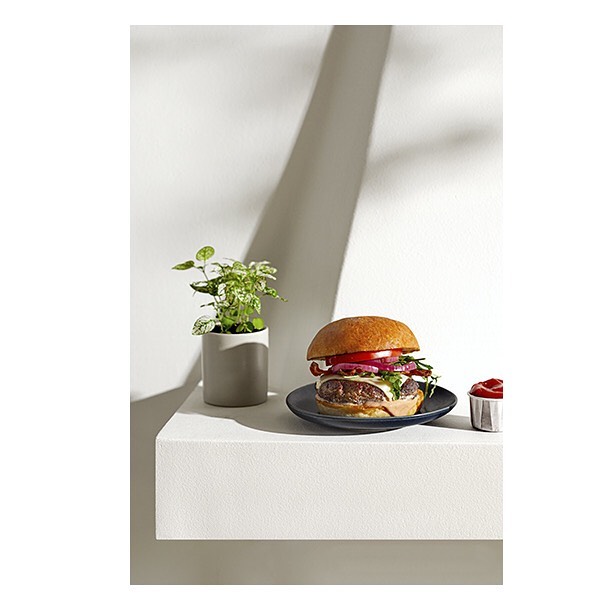 Delicious hamburger with toppings on a dark plate, accompanied by a plant and a small bowl of ketchup on a white shelf.